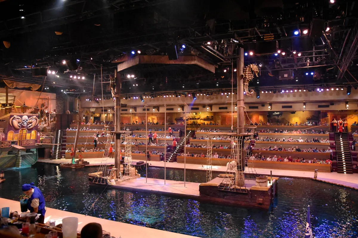 This image presents the vibrant interior of a live performance venue featuring a pirate ship set, a water stage, costumed performers, and an engaged audience. The atmosphere is immersive and dynamic, with children and adults alike enjoying the spectacle.