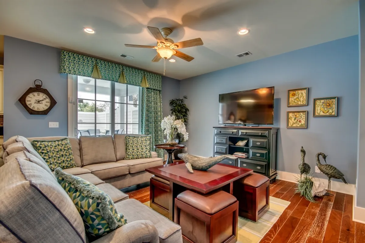 A cozy, welcoming living space featuring a plush sofa with patterned cushions, a coffee table adorned with decorative touches, and a television unit, all bathed in natural light from a large window. The arrangement invites guests to unwind, socialize, or enjoy quiet moments together.