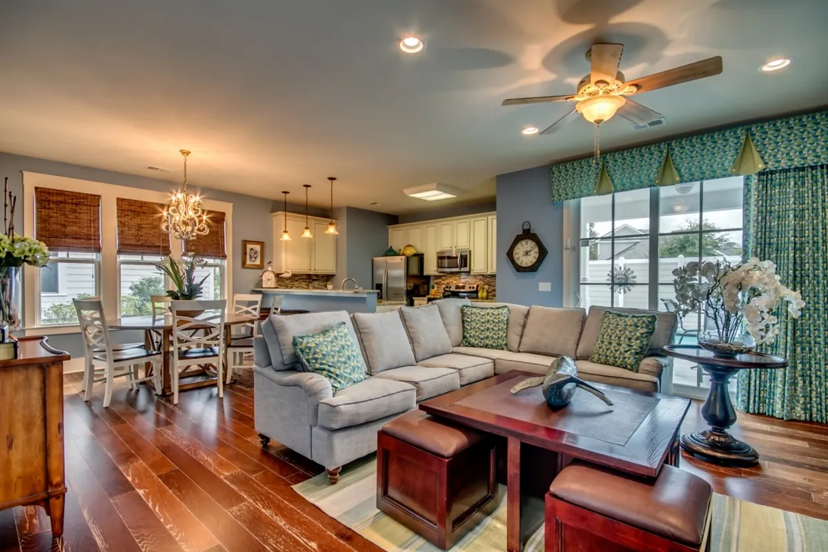 A wide view of the open-concept living area featuring a plush sofa for relaxing, a dining table positioned beneath a stylish chandelier, and the fully equipped kitchen seamlessly integrated into the space. Sunlight streams in through large windows, illuminating the entire area and inviting guests to gather, entertain, or unwind together.