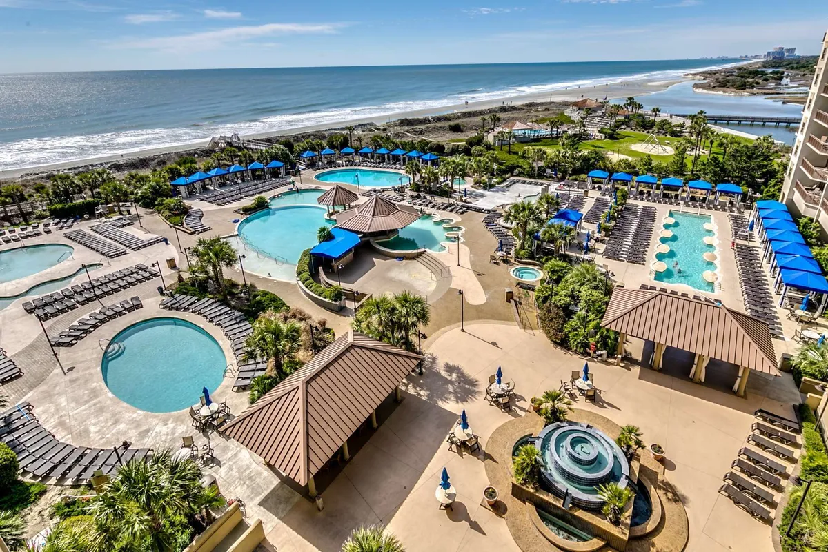 A vibrant resort pool complex featuring several sparkling swimming pools, surrounded by palm trees and dotted with lounge chairs and cabana structures. The scene exudes a vacation-like, serene mood with views extending toward beach access, suggesting a luxurious and expansive outdoor escape.