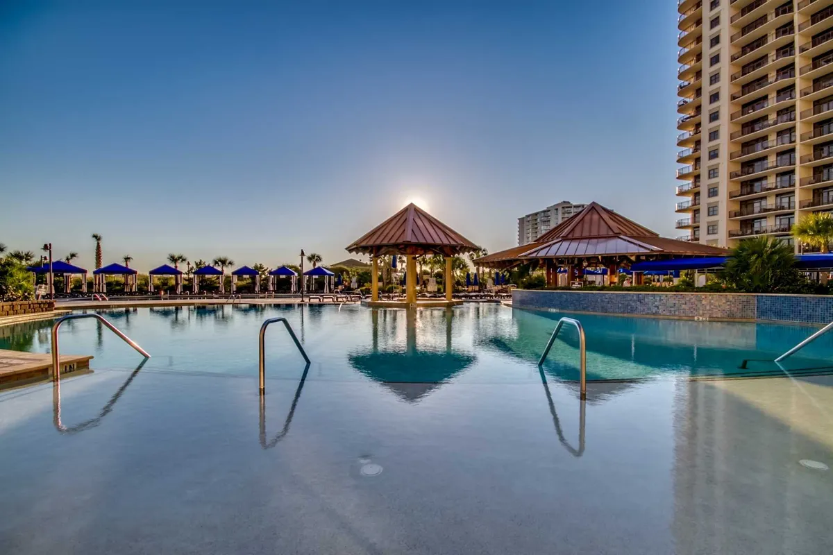 An inviting outdoor swimming pool surrounded by palm trees and stylish cabanas, with the sky transitioning to sunset hues. The tranquil water reflects the soft light, and there are lounge areas perfect for relaxing or socializing. The atmosphere evokes a luxurious, resort-style experience.