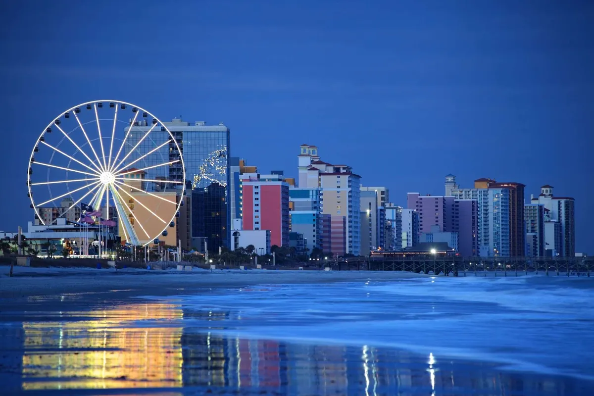 A vibrant view of the local beachfront, with a Ferris wheel, colorful buildings, and gentle ocean waves. The image conveys a lively, festive atmosphere with opportunities for beachside exploration, amusement, and relaxation.