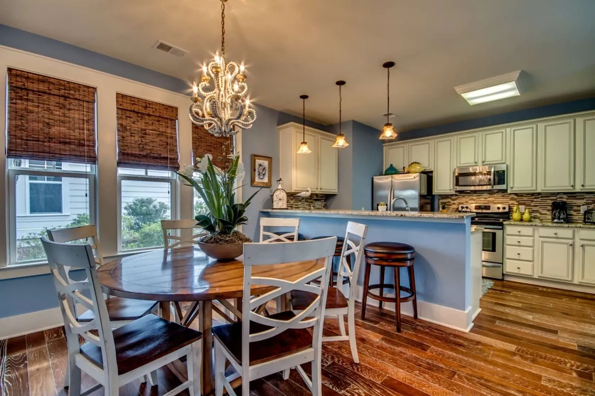 A detailed view of the dining area with a modern chandelier overhead, a spacious dining table ideal for family meals or game nights, a kitchen island in the background for casual gatherings, and windows with blinds letting in soft natural light.