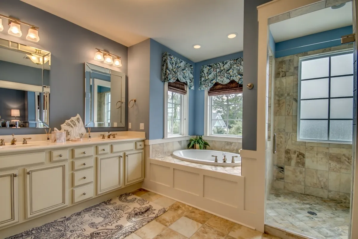 A luxurious bathroom scene with a large bathtub, a separate shower area, a double sink vanity, and windows dressed with soft curtains. The space is bright with natural light and exudes a calming, spa-like feel.