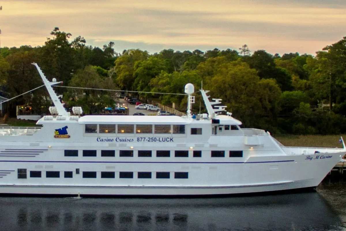 This image features a casino cruise ship docked amid lush greenery and calm waters, capturing guests enjoying activities like dining, socializing, and watching the sunset from the deck.